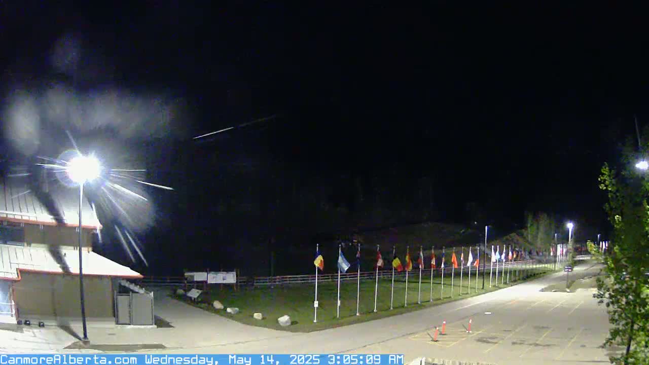 A nighttime scene shows a meteor shower streaking across a dark sky above a paved area with a row of flags and a building.