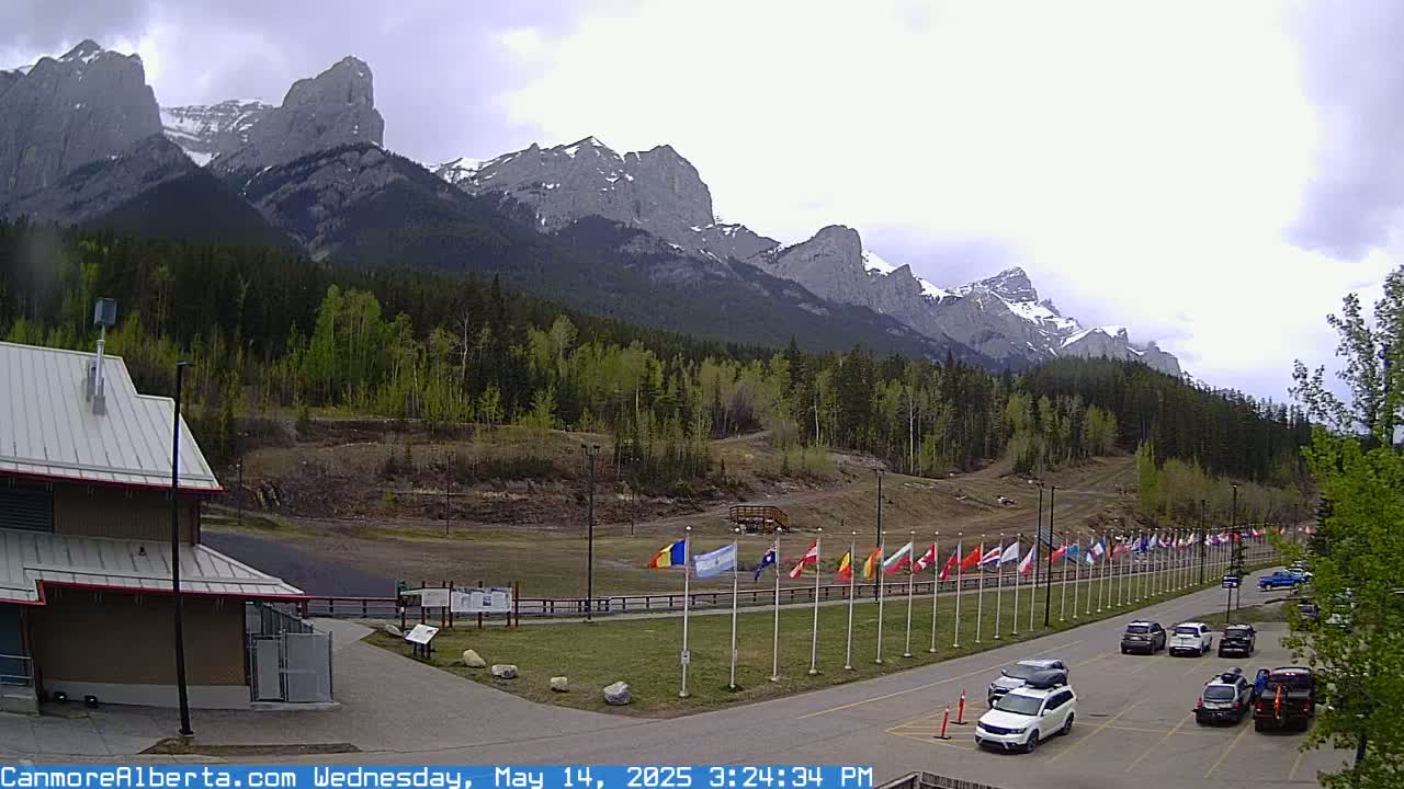 A parking lot with several cars sits before a row of flags displayed against a backdrop of a mountain range under an overcast sky.