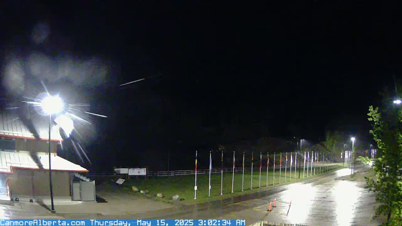 A nighttime, wet scene shows a building, a row of flags, and a road under a dark sky with light rain and several shooting stars.