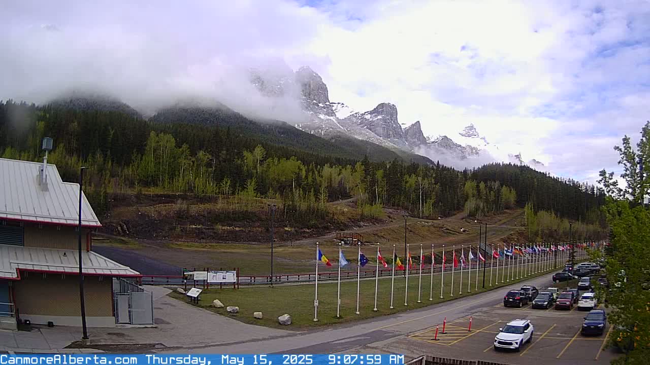 A partially cloudy sky hangs over a mountain range partially obscured by fog, a row of flags, a parking lot with several cars, and a building.