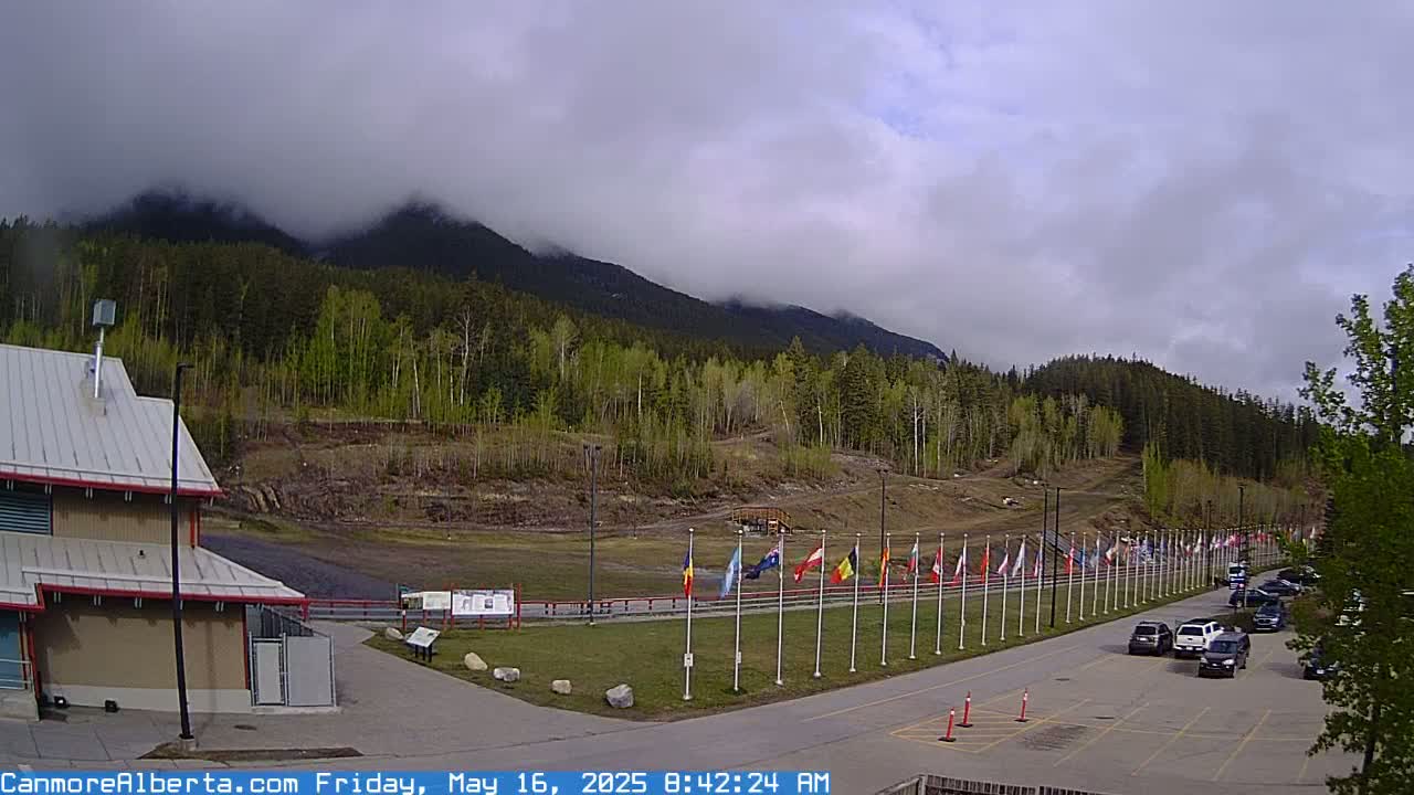 A partly cloudy day reveals a mountain range partially obscured by fog, with a row of flags lining a road next to a building and a small parking lot filled with cars.