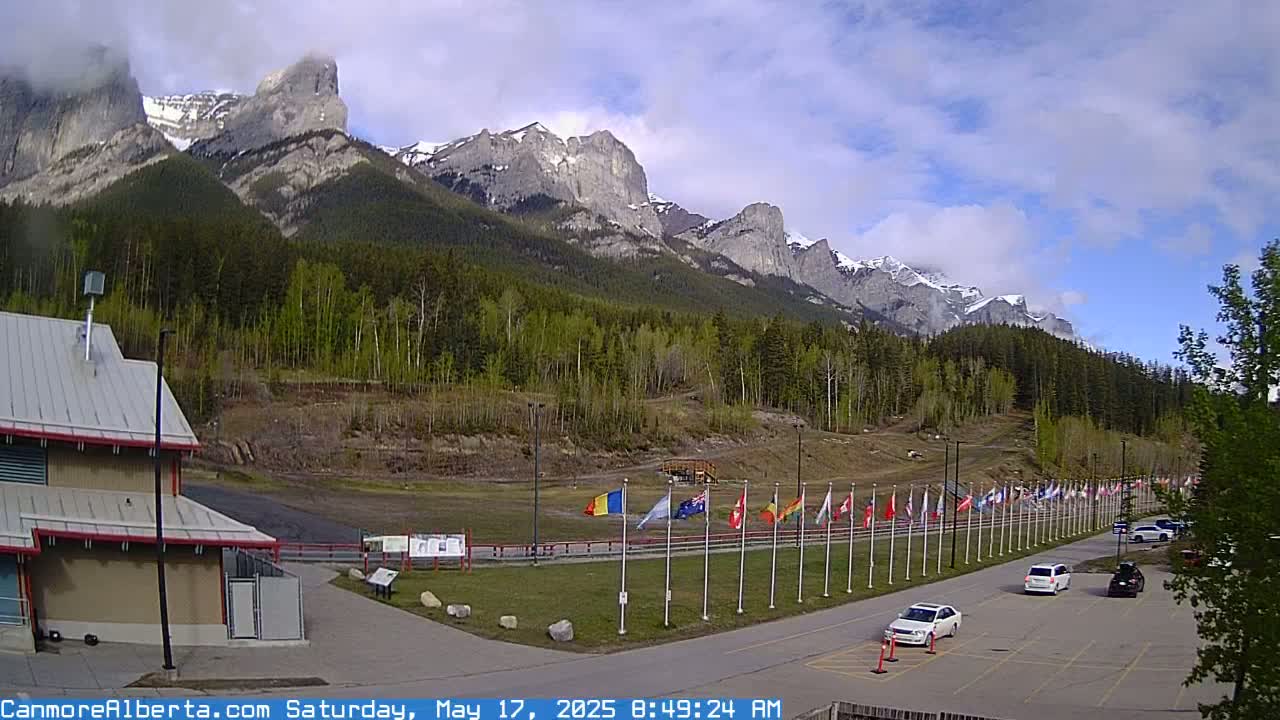 A partly cloudy day shows a parking lot with several cars, a building, and numerous flags displayed in front of a mountainous landscape covered in evergreen trees.