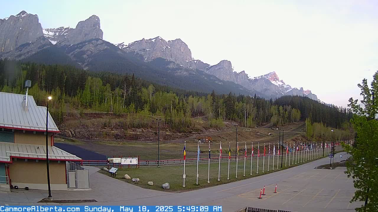 A parking lot and a building sit in the foreground, with a grassy area leading to a row of flags in front of a forest and a mountain range under a clear, bright sky.