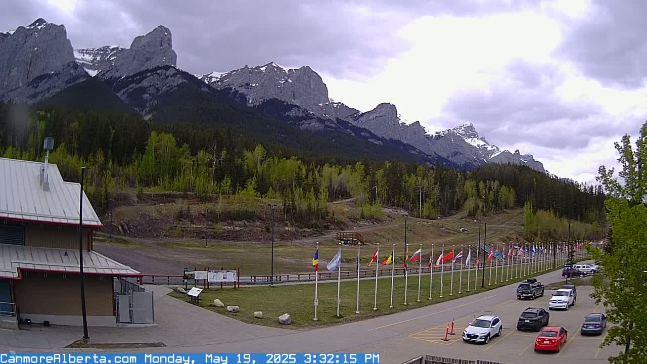 A parking lot with several cars is situated in front of a building, with a row of flags and a mountain range in the background under a cloudy sky.