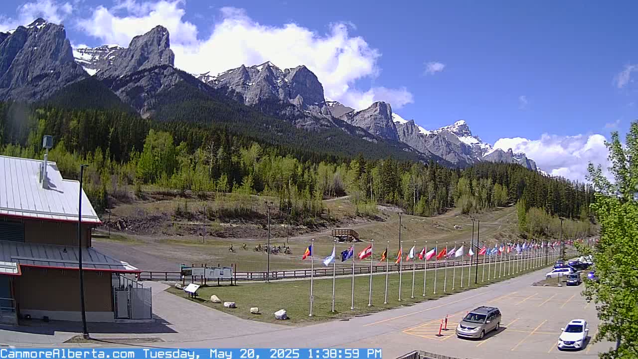 A partly cloudy day reveals a mountainous landscape with a parking lot, several vehicles, and a row of flags in front of a building.
