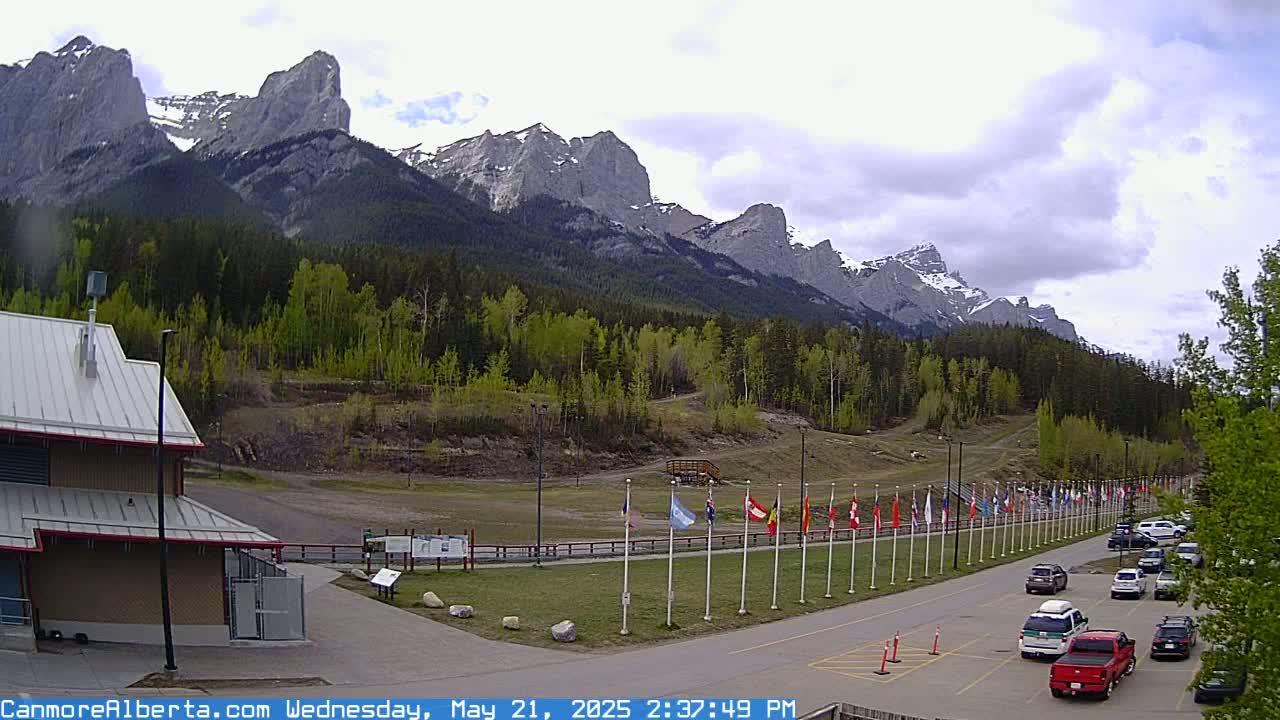 A parking lot with several cars sits before a row of flags in front of a forested mountain range under a partly cloudy sky.