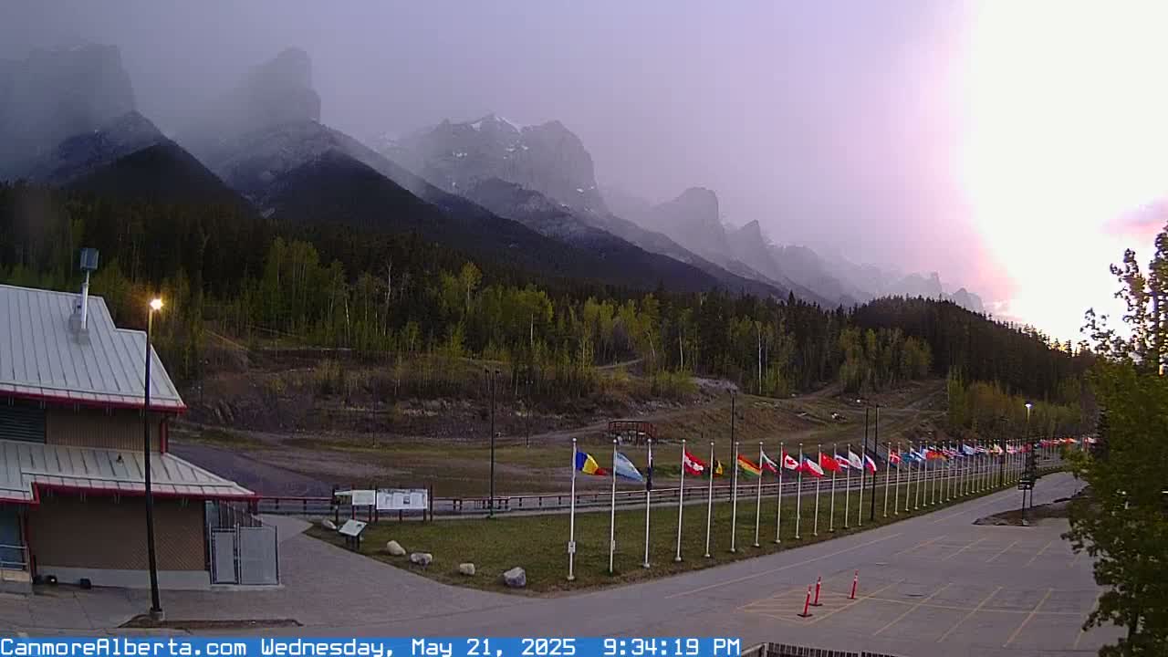 A hazy sunset casts a purple glow over a row of flags situated in front of a mountain range, partially obscured by light rain or mist.