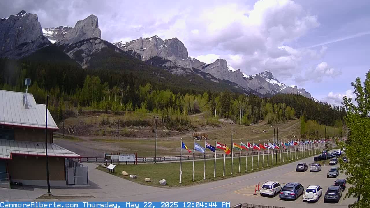 A partly cloudy day shows a parking lot with cars in front of a row of flags, a building, and a mountainous landscape with snow-capped peaks in the background.