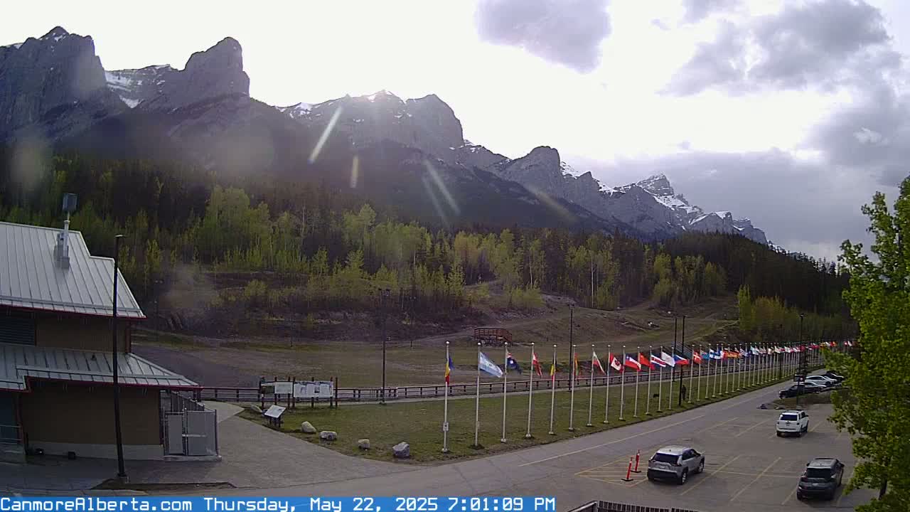 A row of flags stands in a parking lot before a mountain range under a partly cloudy sky.