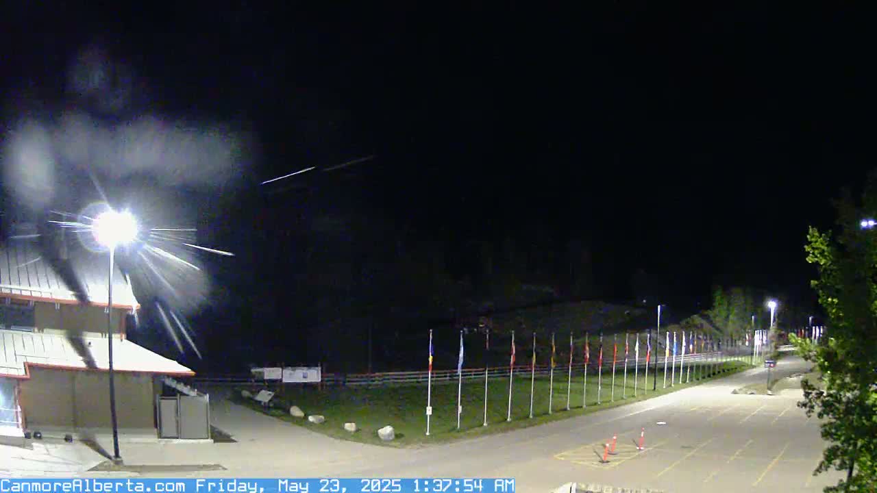 A nighttime scene shows a paved roadway leading to a row of flags, a building to the left, and light streaks in the sky possibly from camera movement or light sources.