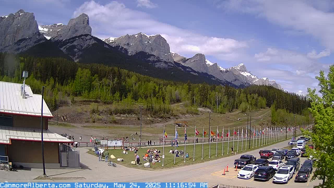 A sunny day reveals a mountain range with snow-capped peaks behind a grassy area where people are gathered, some sitting and others standing, with a parking lot full of cars in the foreground and a building to the left.