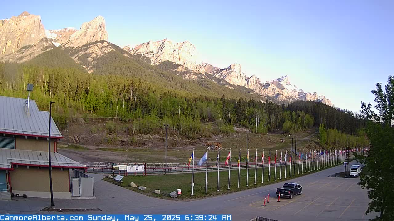 A clear morning reveals a parking lot with a few vehicles, a line of flags, and a building in the foreground, all set against a backdrop of a mountain range covered in lush green forests.