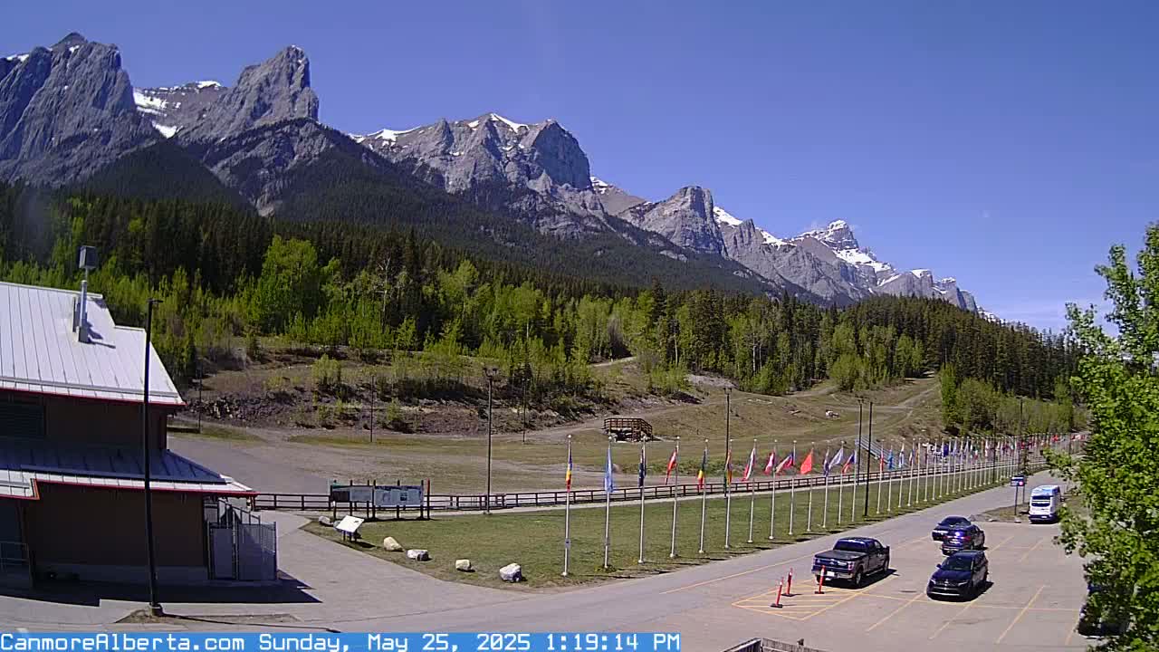 A parking lot with several cars is situated in front of a building, with a row of flags in the mid-ground, all set against a backdrop of a mountain range under a clear, sunny sky.