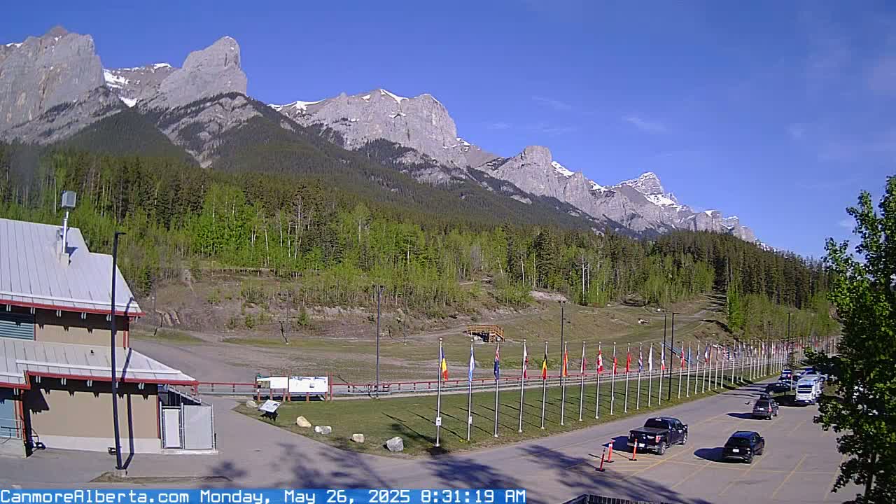 A parking lot with several cars sits before a row of flags in front of a grassy area and a forested mountain range under a clear, sunny sky.