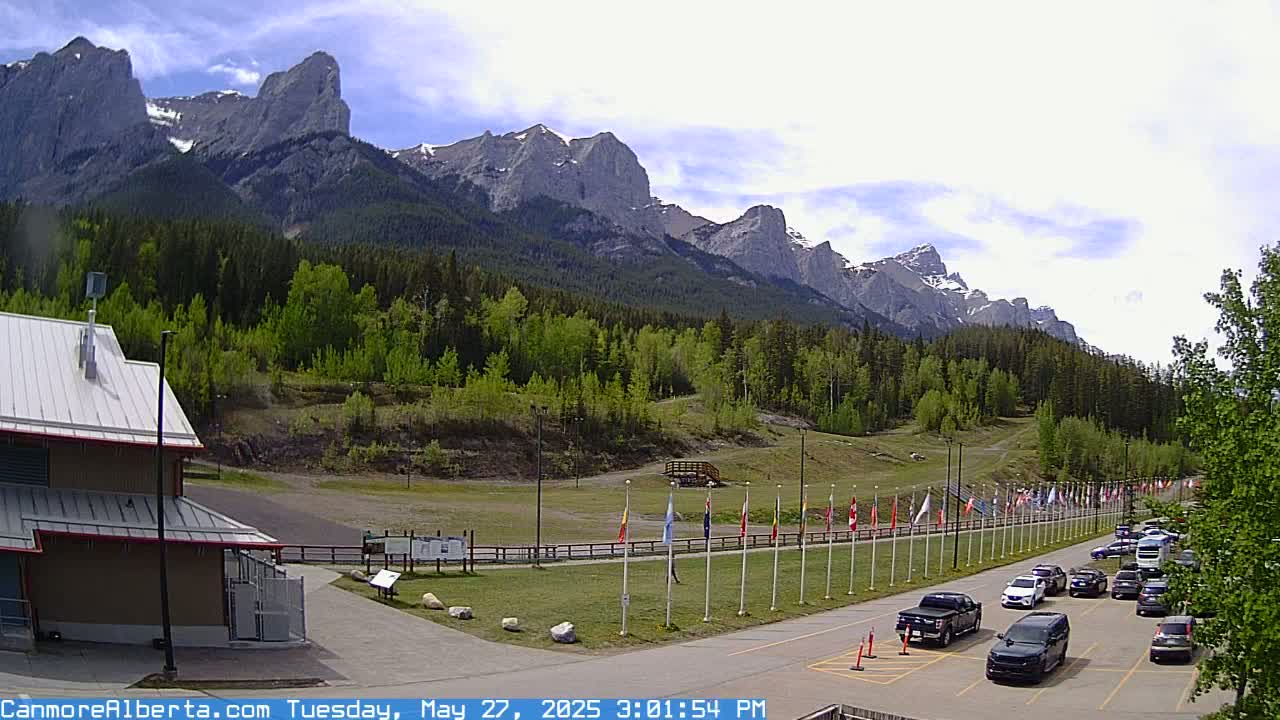 A parking lot with cars is situated in front of a row of flags, a grassy area, and a forested mountain range under a partly cloudy sky.
