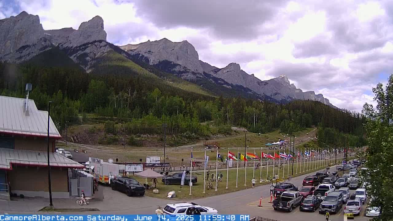 A partly cloudy day reveals a mountain range behind a parking lot filled with cars, a few people, and various flags lined up near a building.