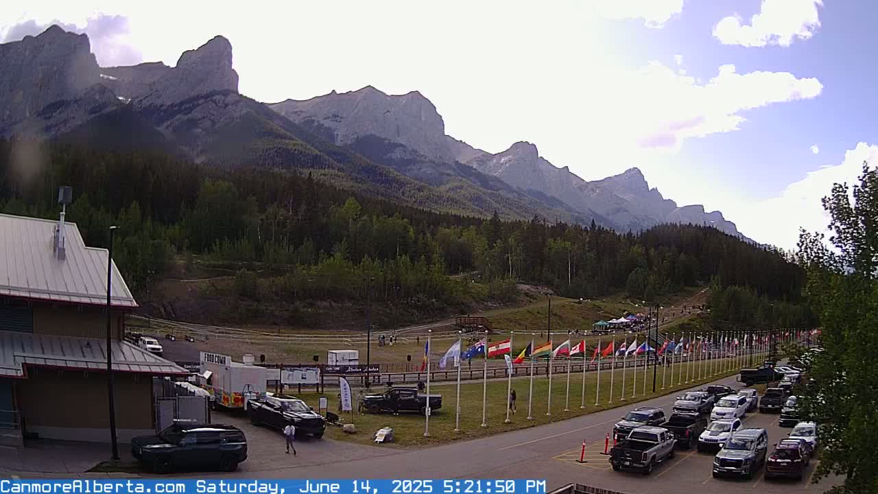 A partly cloudy day reveals a mountain range behind a parking lot filled with cars, a few people, and various flags lined up near a building.