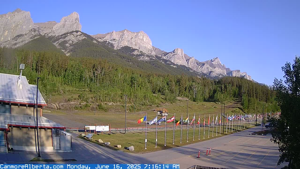 A row of flags stands in a parking lot before a forested mountain range under a clear blue sky.