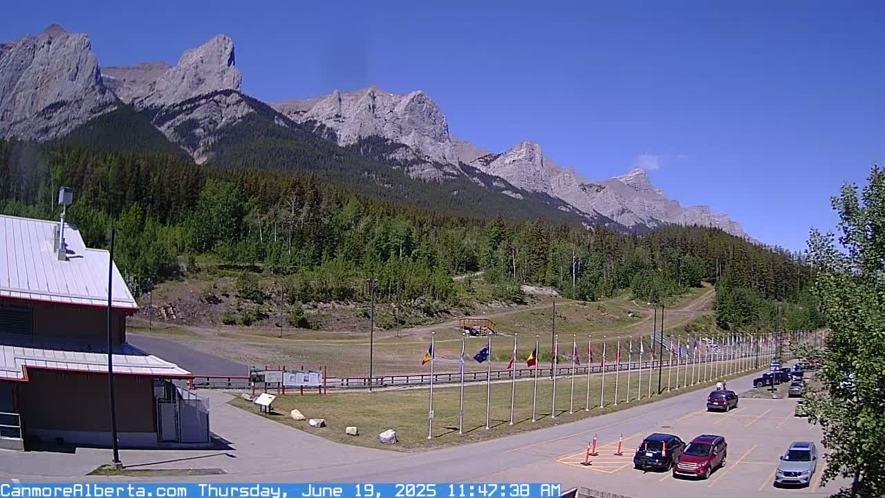 A parking lot with several cars is situated in front of a building near a grassy area with flags, all set against a backdrop of mountains under a clear blue sky.