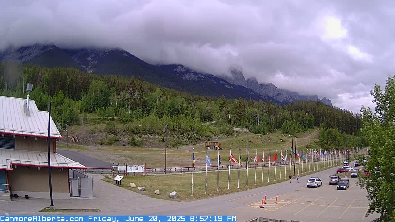 A cloudy day reveals a mountainous landscape with a row of flags lining a paved area next to a building, a few cars parked nearby, and some pedestrians walking.
