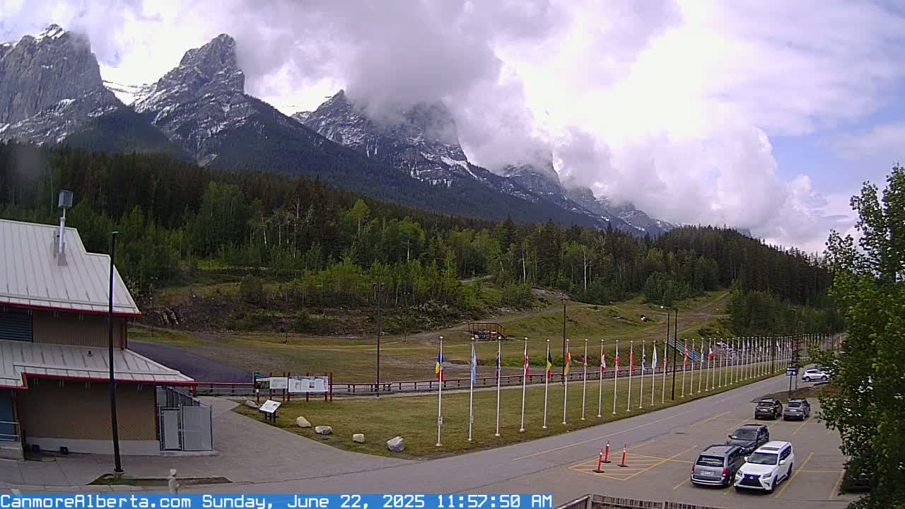 A partly cloudy day shows a parking lot with several cars, a row of flags, and a building at the base of a snow-capped mountain range covered with a lush green forest.