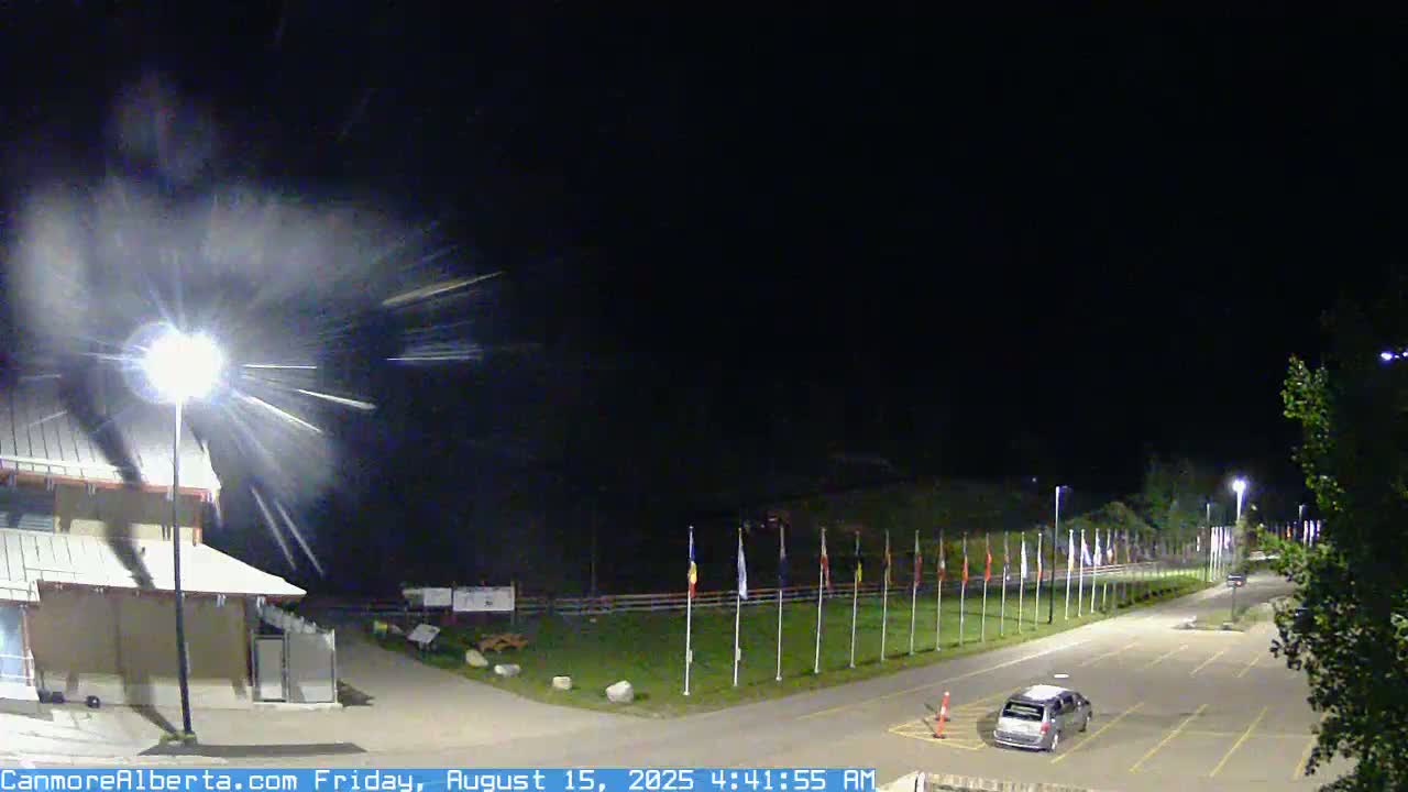 A nighttime scene shows a building, a parking lot with a few cars, and a line of flags under a dark sky with light snow or dust falling.