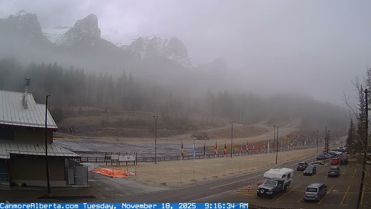 A nighttime scene shows a building, a parking lot with a few cars, and a line of flags under a dark sky with light snow or dust falling.