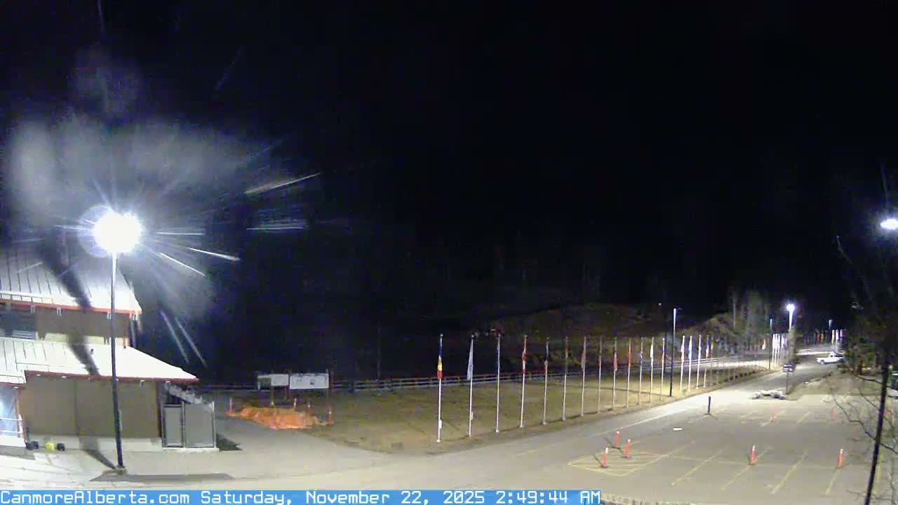 Under snowy nighttime conditions, a brightly illuminated building stands adjacent to a parking lot and road lined with multiple flagpoles and streetlights, extending towards distant trees.