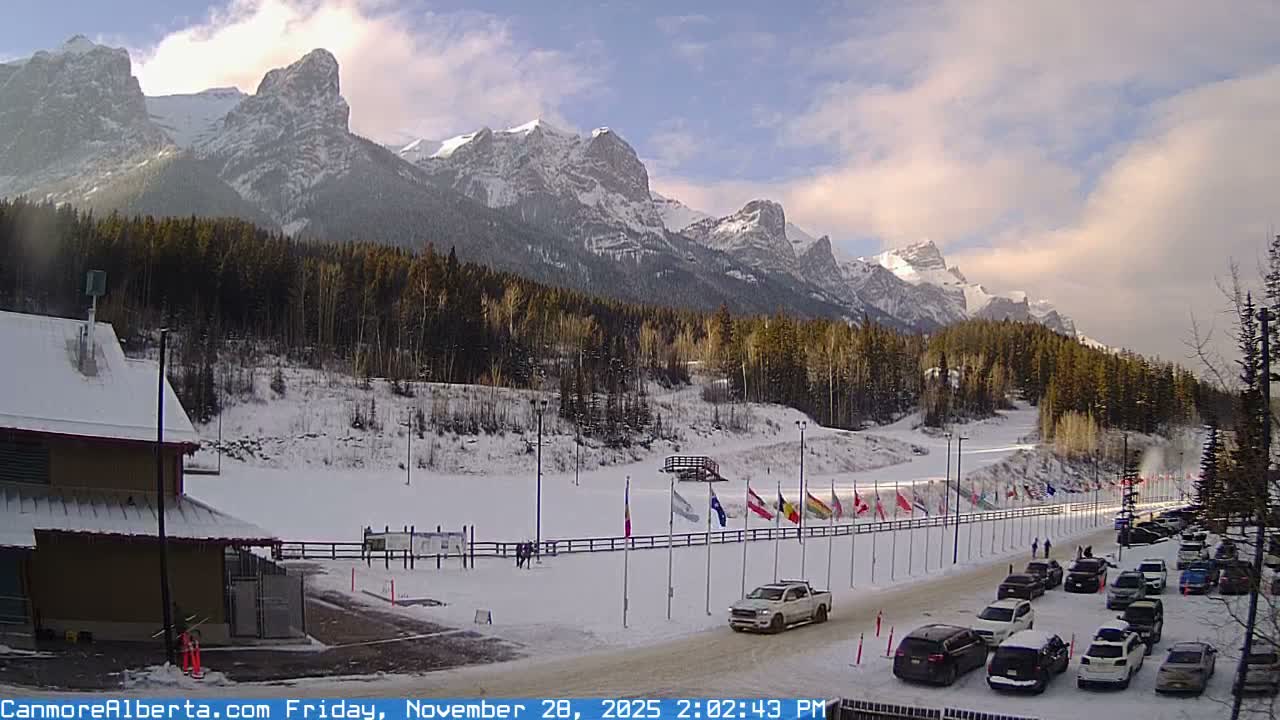 A snowy mountain landscape featuring rugged, snow-capped peaks under a partly cloudy sky, with evergreen forests, a building on the left, a road with parked cars, and a long line of flags lining a snow-covered field.