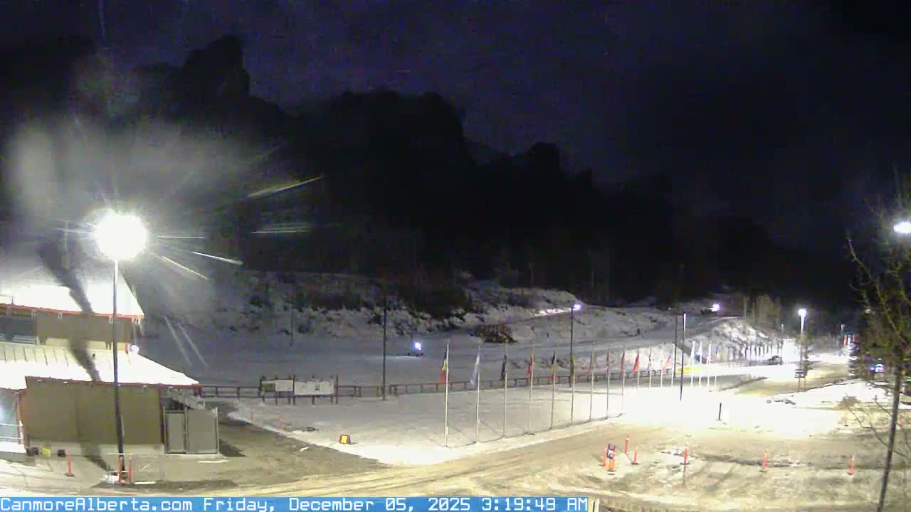 A clear, cold, and snowy night illuminates an outdoor winter scene with artificial lights highlighting snow-covered grounds, a building, and flagpoles against a backdrop of dark mountain silhouettes.