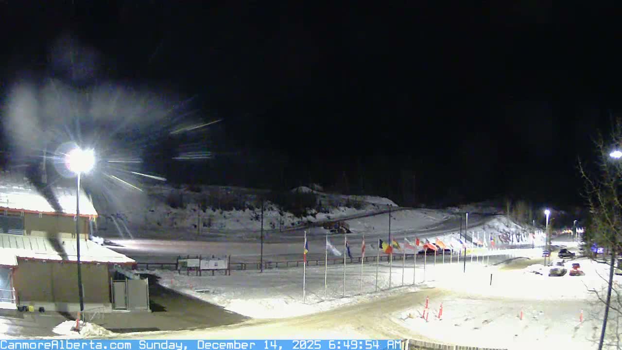 A clear, cold, and snowy night illuminates an outdoor winter scene with artificial lights highlighting snow-covered grounds, a building, and flagpoles against a backdrop of dark mountain silhouettes.