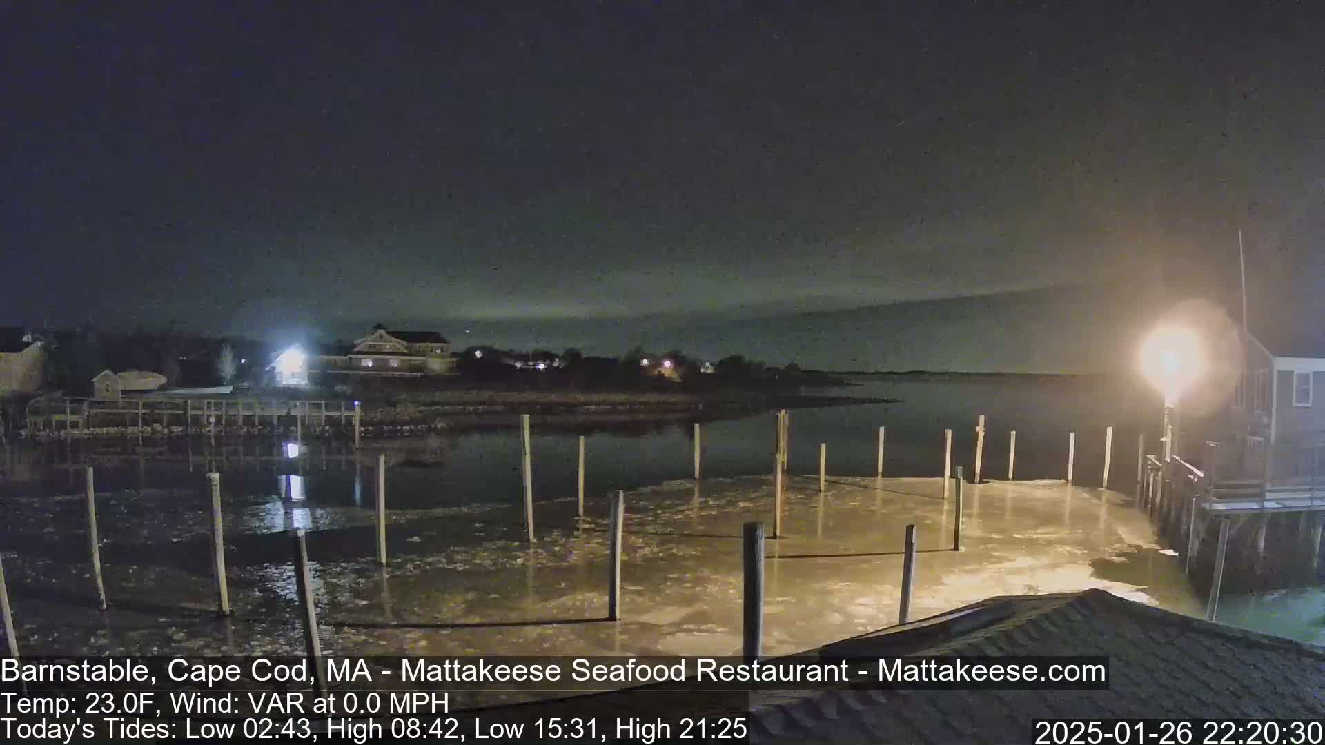 A nighttime view of a partially ice-covered dock and calm water, with buildings and lights visible in the background under a clear, dark sky.