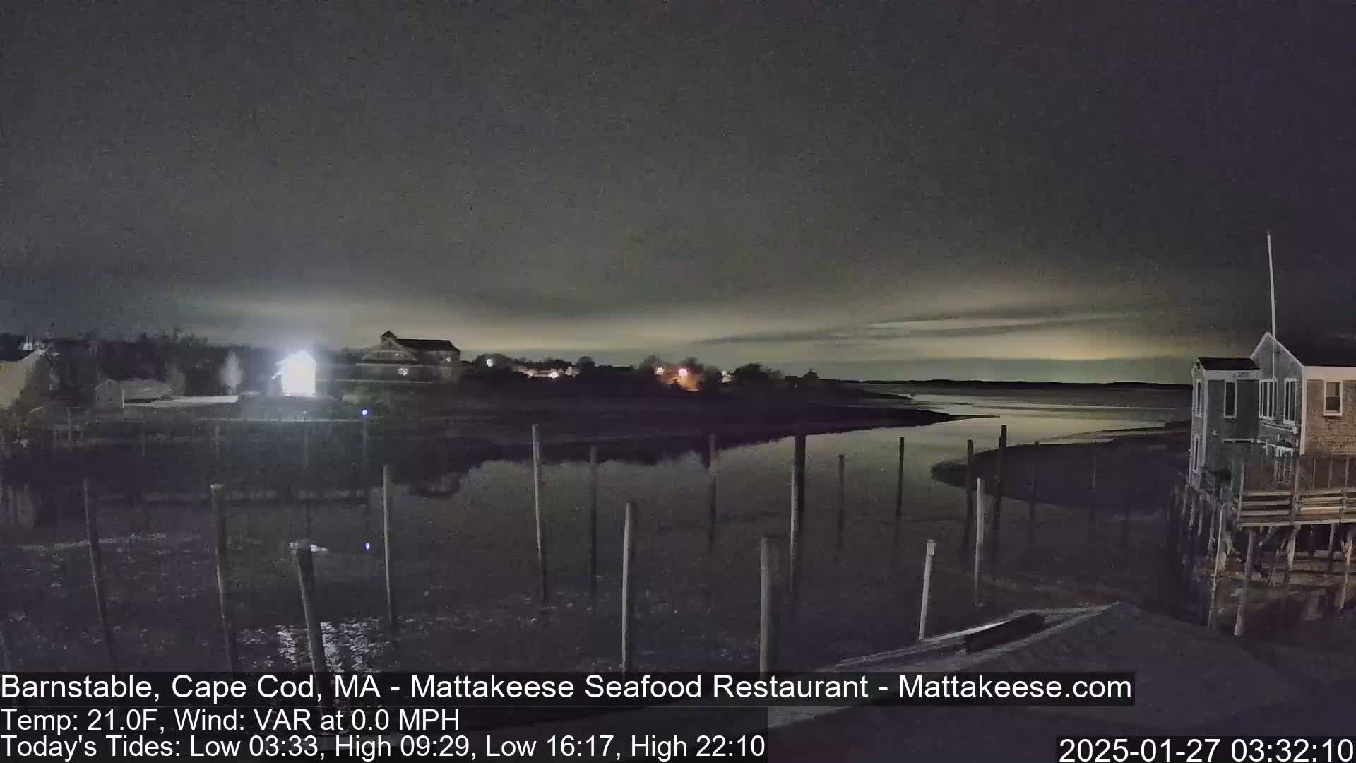 A nighttime view of a calm body of water with a shoreline featuring buildings and pilings under a dark, mostly clear sky.