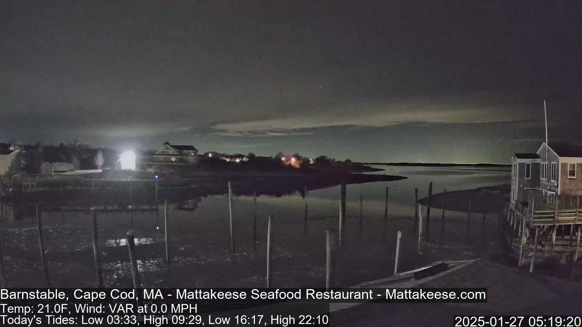 A nighttime view of a calm body of water with a few buildings and pilings visible under a dark, mostly clear sky.