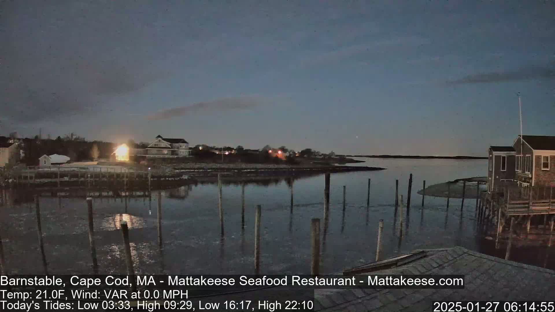 Under a twilight sky, a calm body of water reflects the lights of houses and buildings along its edge, with wooden pilings extending into the water.