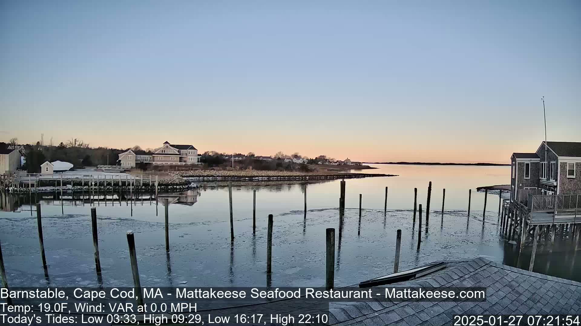 A calm, twilight scene over a partially frozen harbor, with houses and a restaurant visible along the shoreline.