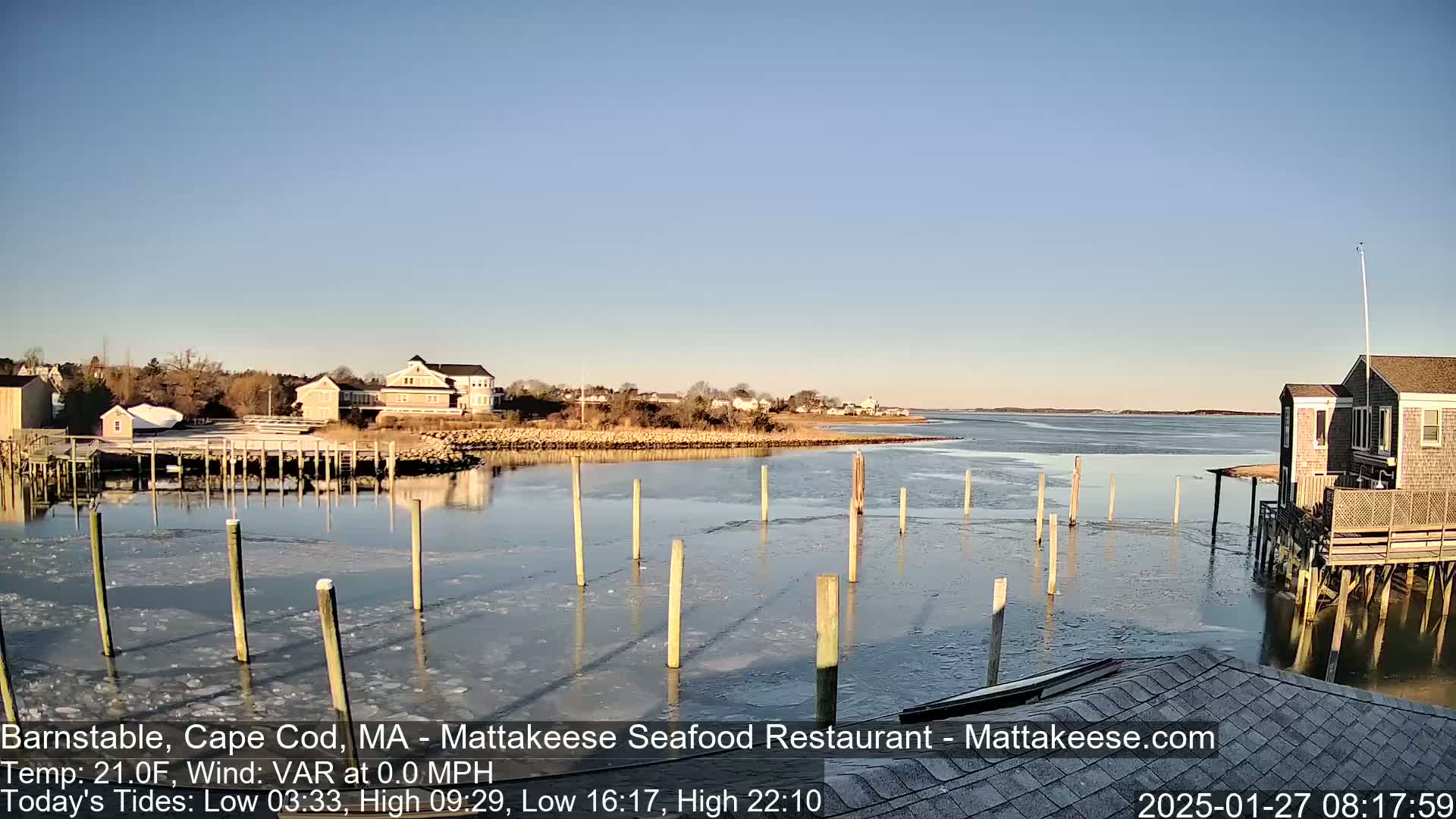 A partially frozen harbor with wooden pilings and buildings on the shore under a clear blue sky.