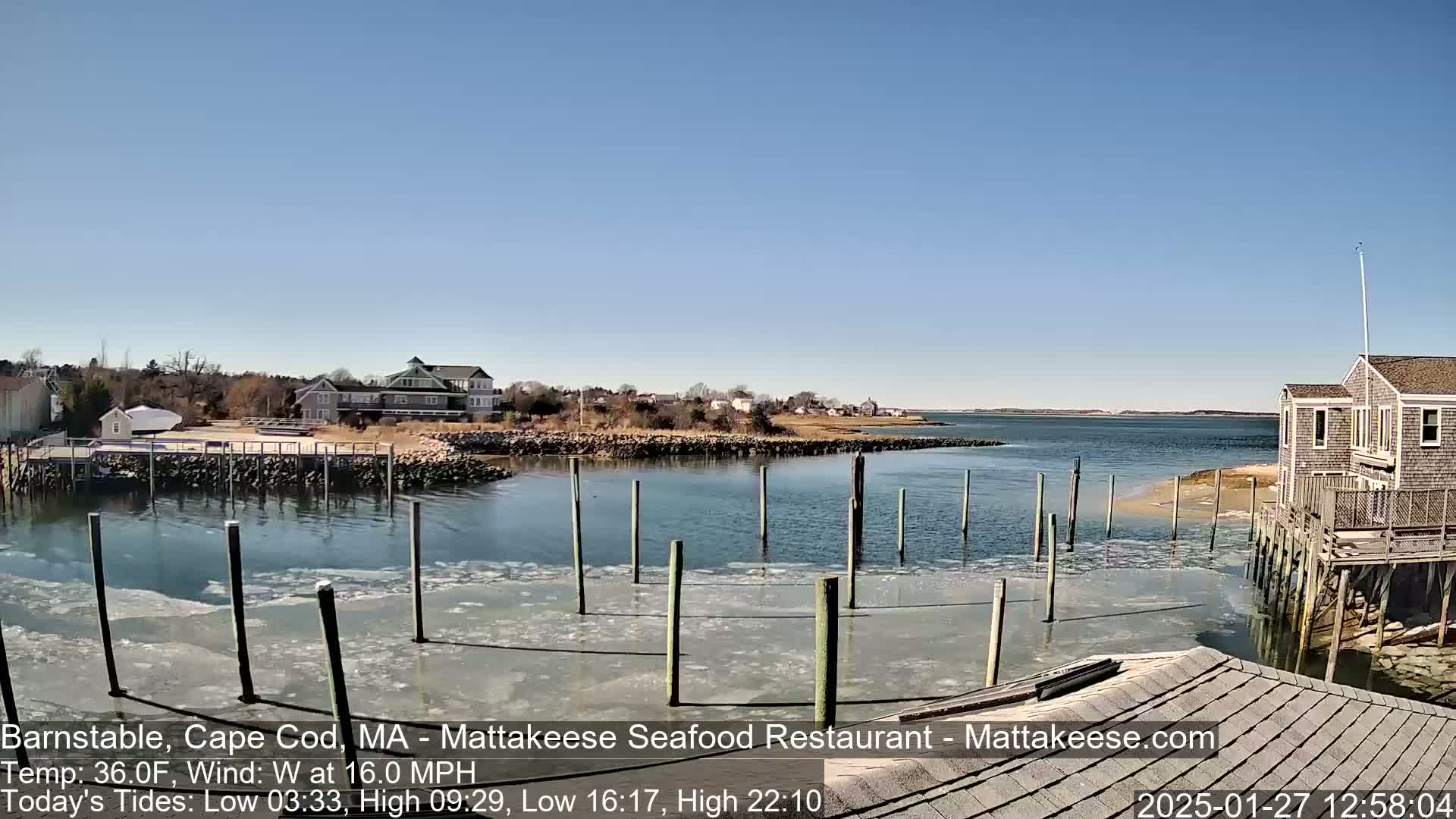 A partly frozen harbor with wooden pilings and houses on the shore under a clear blue sky.