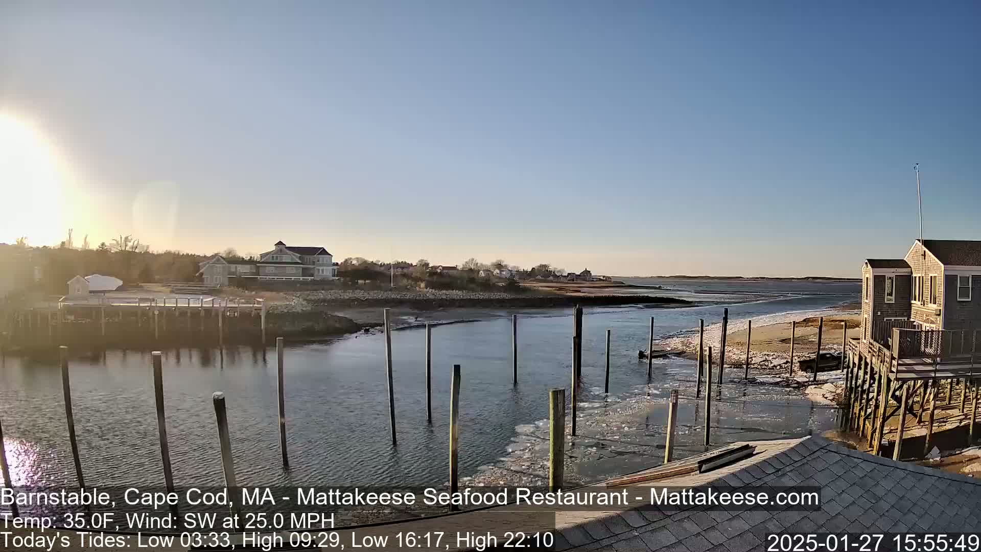 A calm, sunny waterfront scene with houses, a dock partially covered in ice, and a sandy shore under a clear sky.