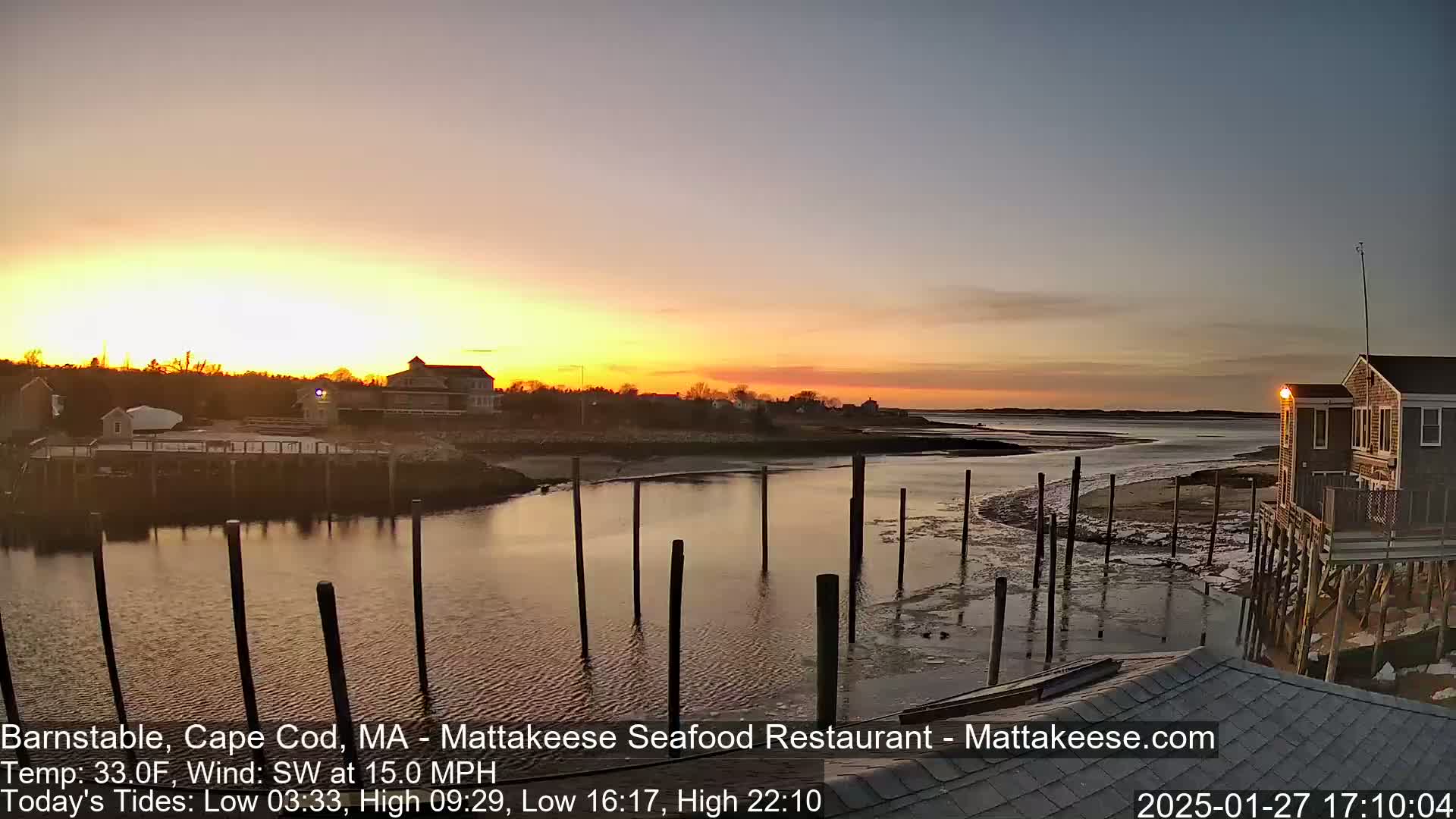 A sunset over calm water with a few buildings and wooden pilings in the foreground.
