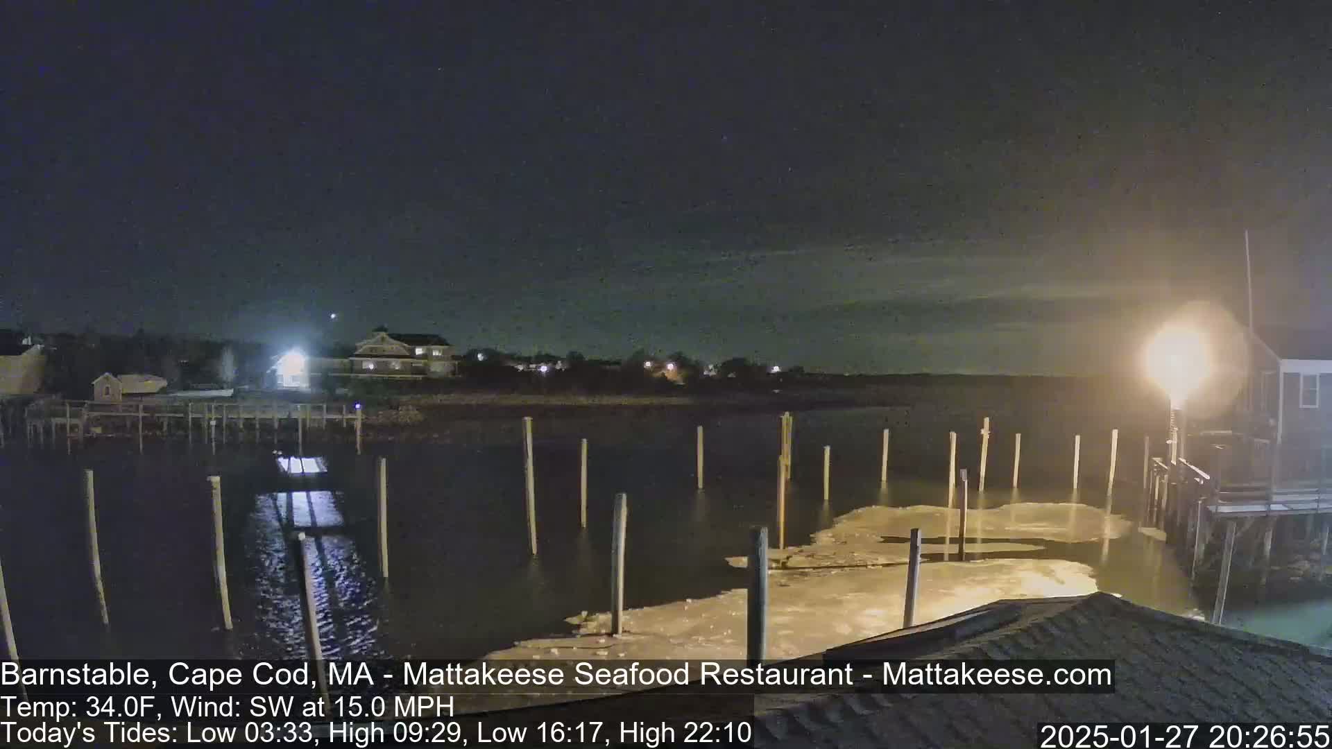 A nighttime view of a calm harbor with wooden pilings, illuminated by lights from nearby buildings under a clear, dark sky.