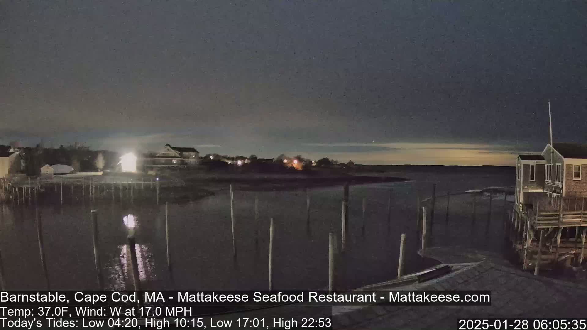 A nighttime view of a calm body of water with wooden pilings, several buildings, and a slightly brightening sky.