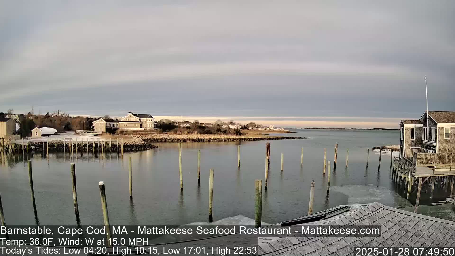 A calm, overcast day shows a waterfront scene with several buildings, a small harbor with pilings in the water, and some ice along the shore.