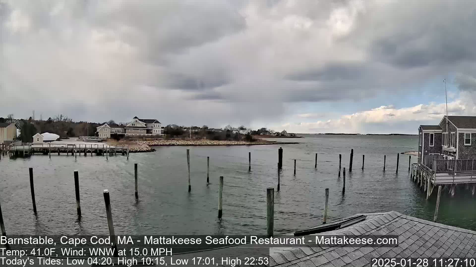 A calm body of water with wooden pilings extends from a shoreline with houses and a cloudy sky overhead.