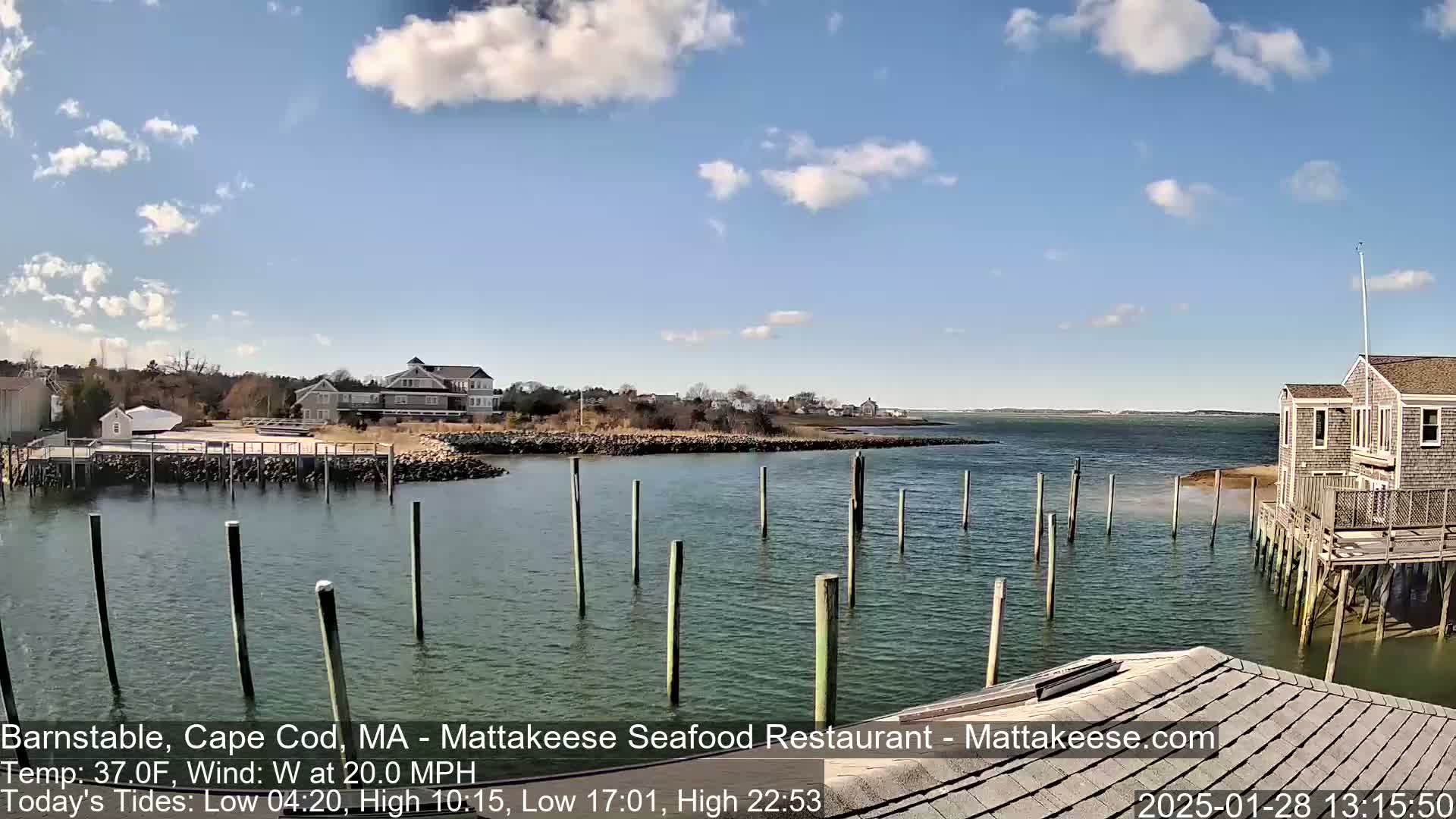 A partly cloudy day with a 20 mph west wind shows a calm harbor with several pilings and a building on stilts, across from a shoreline with buildings and trees.