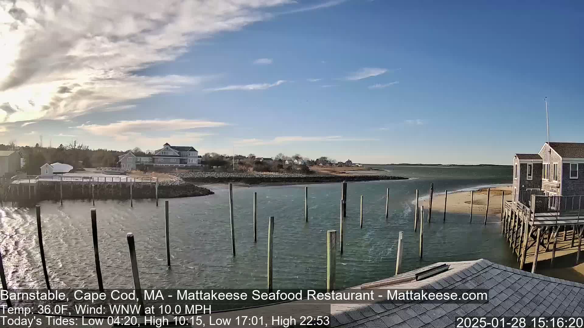 A partly sunny day with a WNW wind at 10.0 mph shows a coastal scene with a calm body of water, wooden pilings, buildings, and a sandy shore.