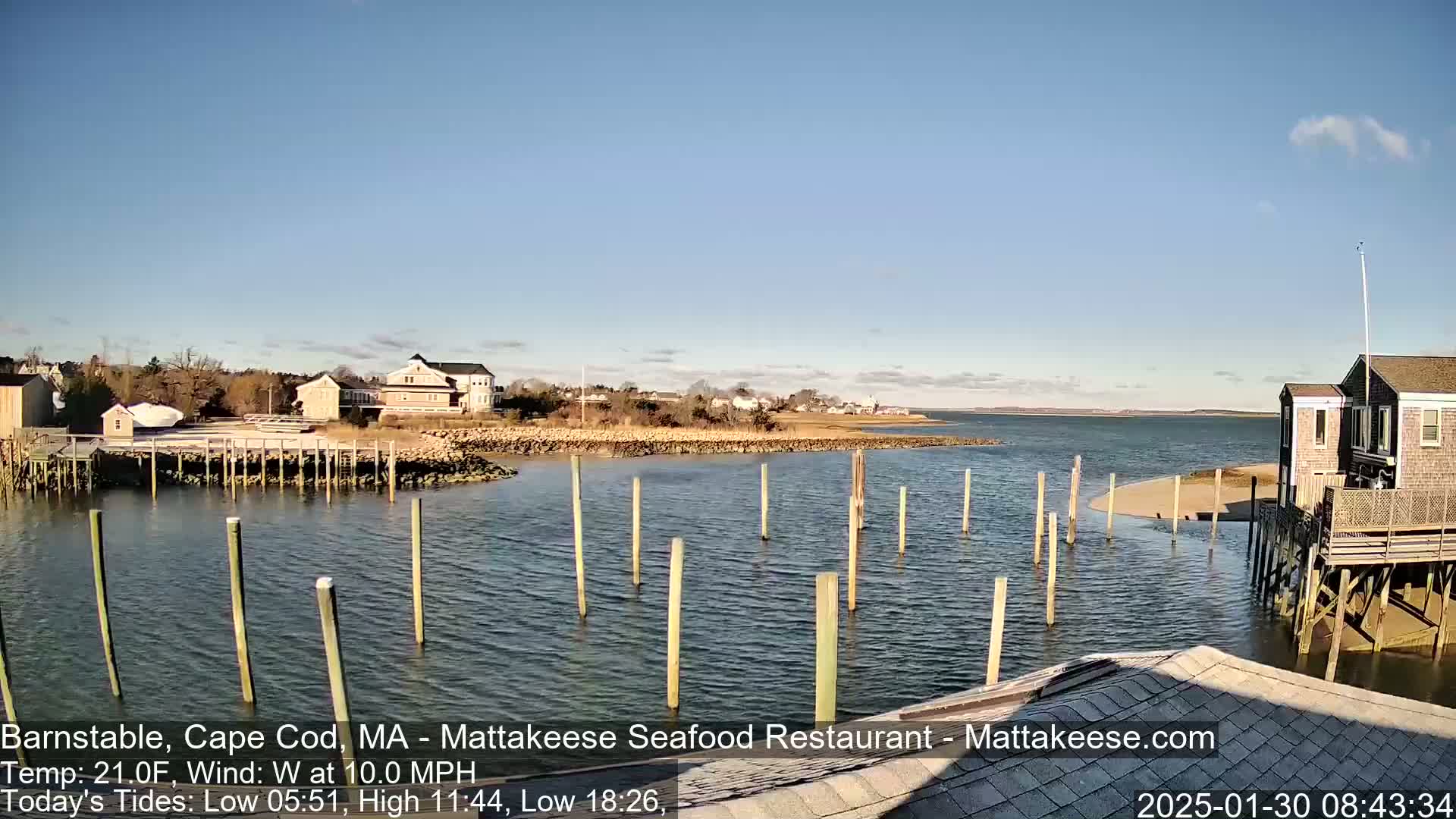 A calm body of water with wooden pilings in the foreground, a small sandy beach, and buildings along the shoreline under a clear, sunny sky.
