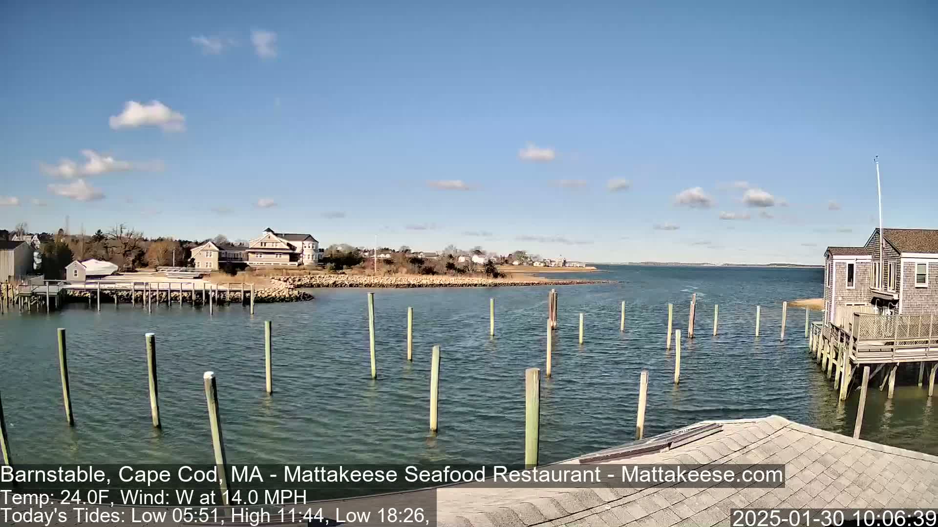 A calm body of water with numerous pilings extends to a shoreline featuring buildings and a partly cloudy sky.