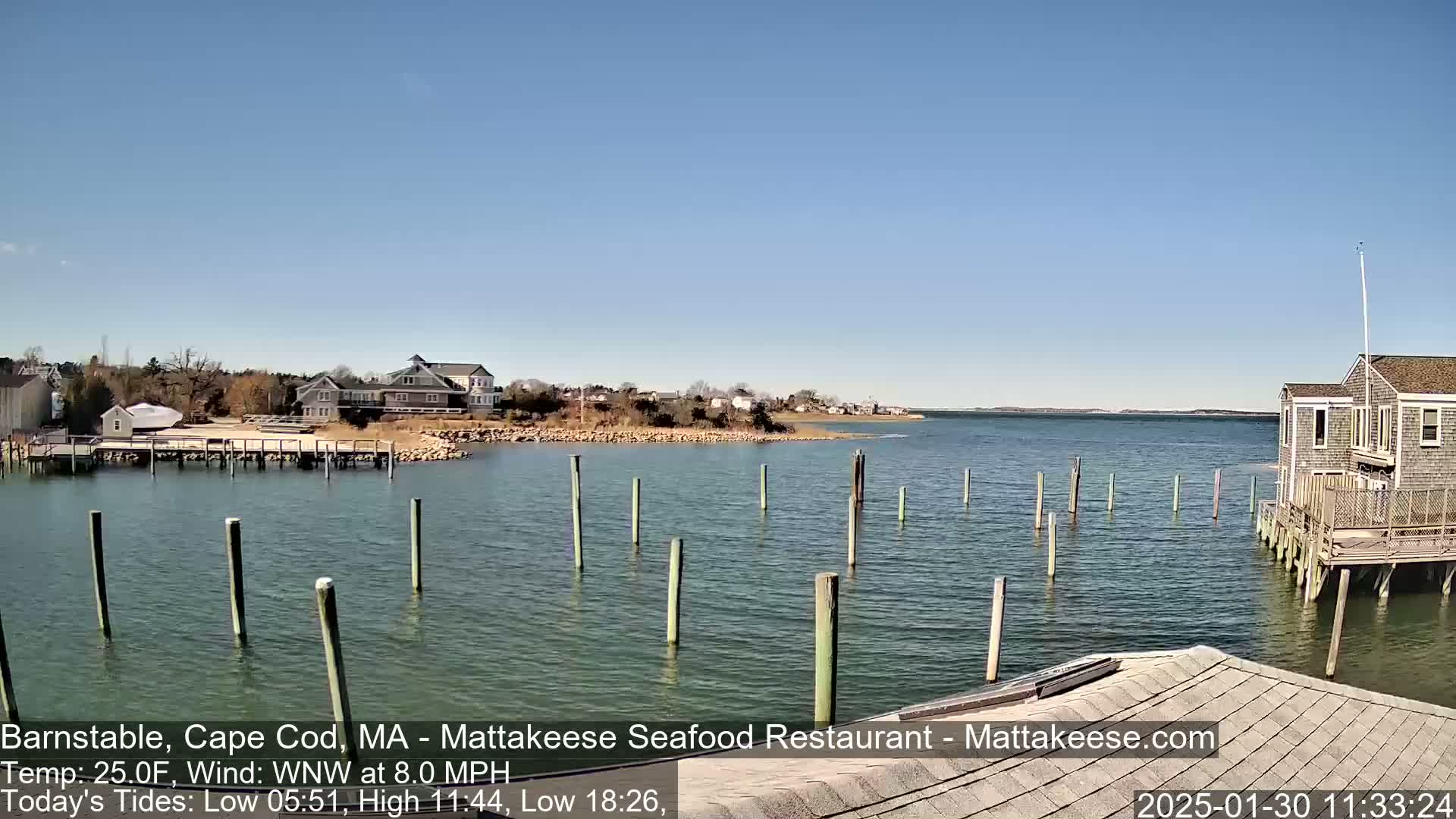 A calm body of water with wooden pilings extends from a shoreline with buildings, under a clear blue sky.