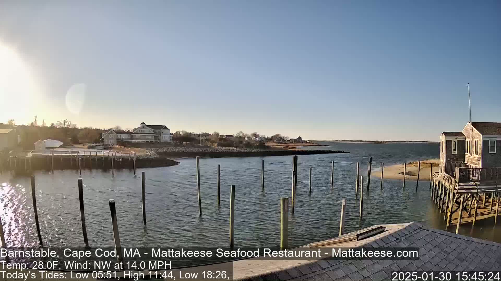 A calm body of water with wooden pilings extends from a shoreline with houses under a clear, sunny sky.